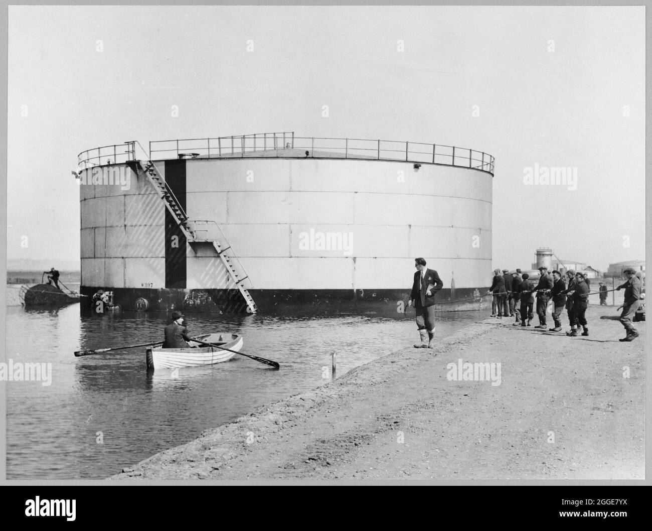 Arbeiter, die während des Baus der Coryton Ölraffinerie einen schwimmenden Benzintank bewegen und den Tank kurz vor dem Abbiegen zeigen. Die Beschriftung unter diesem Albumdruck lautet „Schwimmende Benzintanks. Coryton. „Admirals Gig“. Zeigt den Tank kurz vor dem Kurvendrehen war das Schlauchboot dafür da, von einer Bank zur anderen zu fahren und die Transportseile zu binden.“ Die Ölraffinerie Coryton wurde im Mai 1954 von Ihrer Majestät Königin Elizabeth, der Königin Mutter, feierlich eröffnet. Es wurde von Vacuum Oil Company betrieben, einer Einheit von Mobil. Ab 1996 wurde die Raffinerie von BP als Teil eines Joint Ventu betrieben Stockfoto