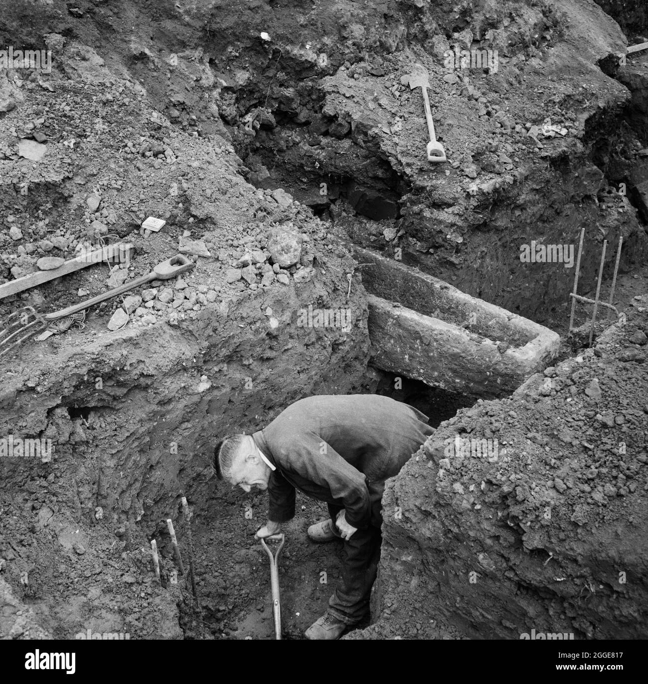 Ein Mann, der in den Pfahlkappengraben auf der Baustelle der Coventry Cathedral grabt, mit einem steinernen Sarg hinter ihm, der in den Graben ragt. Dieses Foto wurde während einer frühen Bauphase der Kathedrale von Coventry aufgenommen, die Abriss, Räumung, Aushub und Pfahlfahren beinhaltete, wobei die ersten Fundamente am Montag, den 7. März 1955, gelegt wurden. Stockfoto