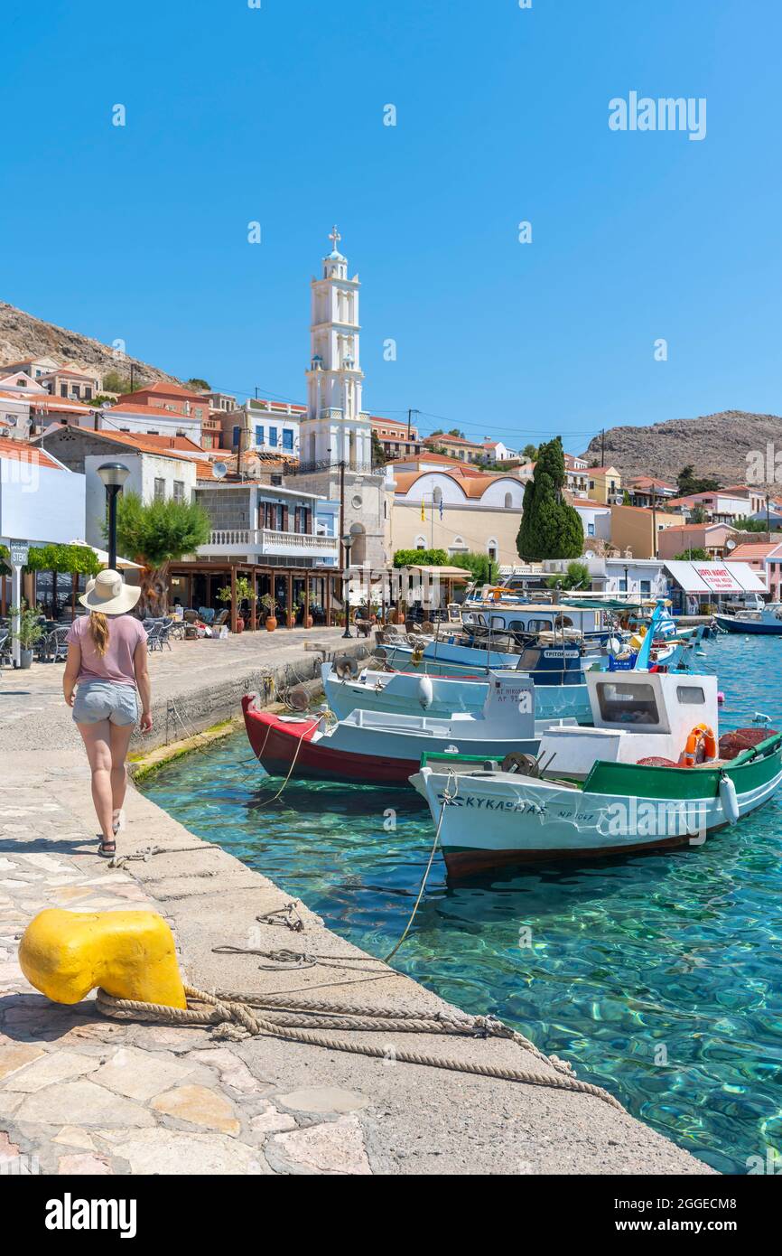 Tourist mit Sonnenhut, Fischerboote in Halki Hafen mit türkisfarbenem Wasser, Promenade mit bunten Häusern von Halki Dorf, Halki, Dodekanes, Griechenland Stockfoto