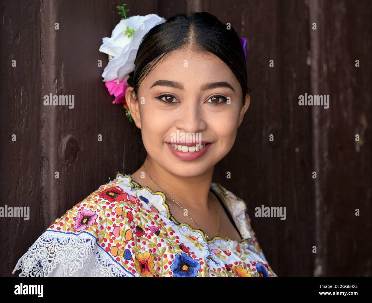 Junge schöne mexikanische Frau mit Make-up trägt traditionelles Maya Yucatecan folklorisches Kleid mit Blumen im Haar und lächelt für den Betrachter. Stockfoto