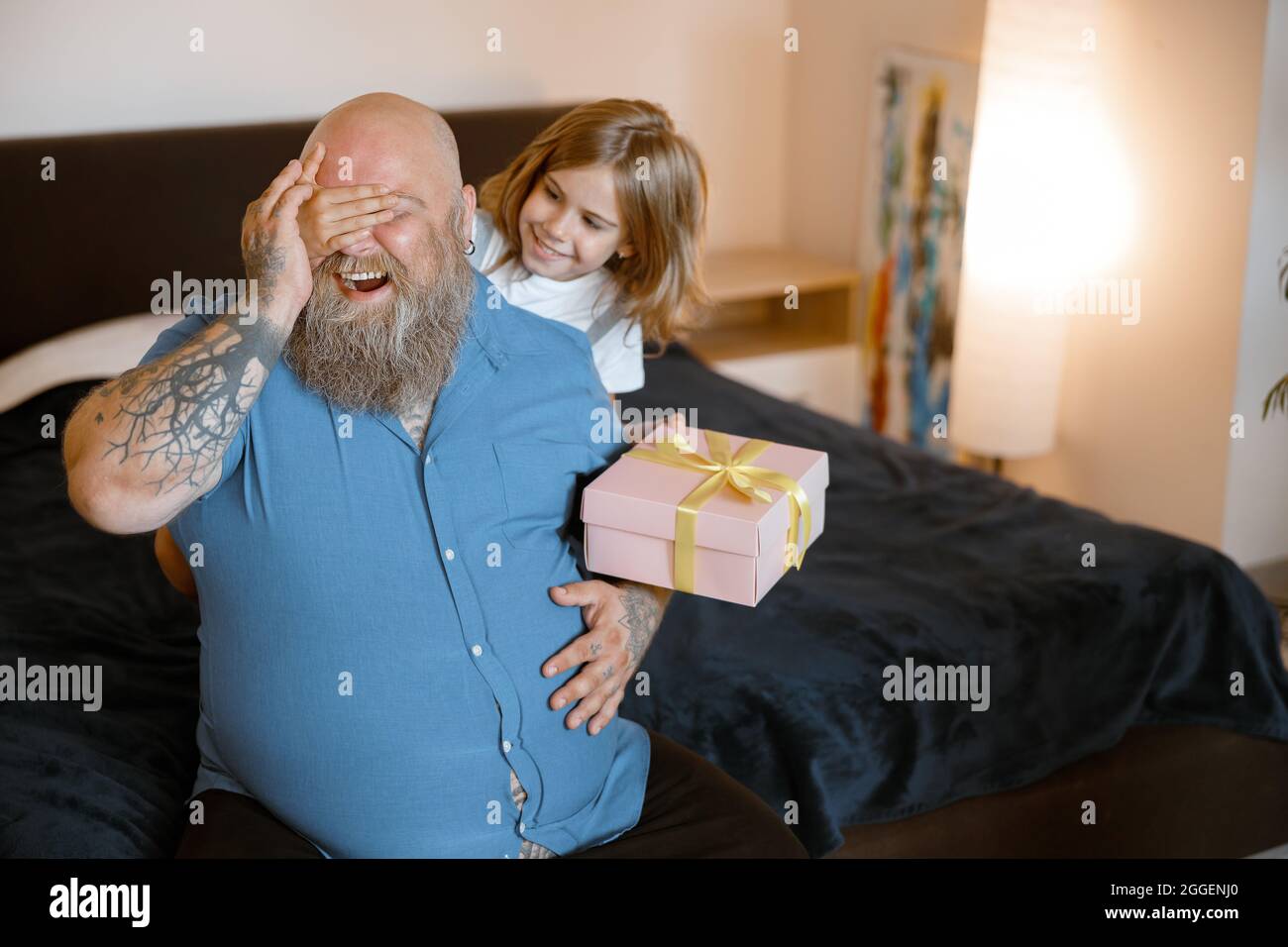 Kleines Mädchen mit Geschenk schließt die Augen des Vaters, um im Urlaub im Schlafzimmer zu überraschen Stockfoto