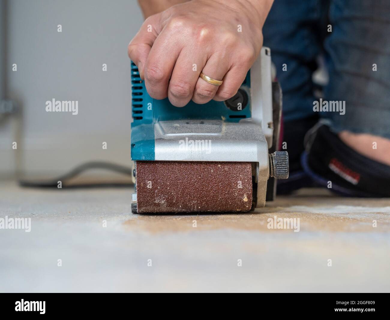 Nahaufnahme einer kaukasischen männlichen Hand, die einen Bandschleifer bediente, um weiße Farbe von einem Holzboden zu entfernen. Stockfoto