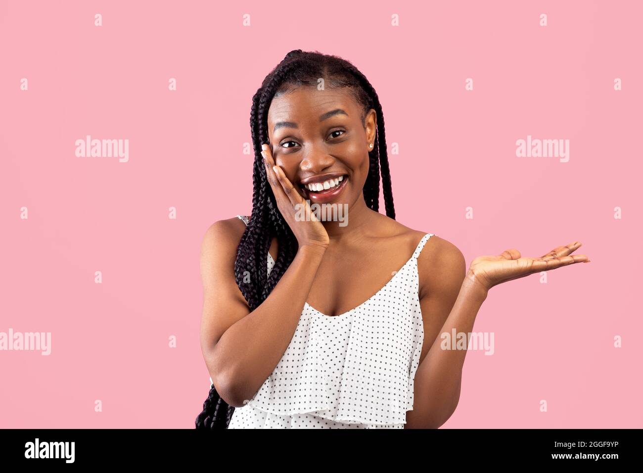 Staunende schwarze Dame berührt ihr Gesicht und zeigt etwas über rosa Studio Hintergrund. Charmante afroamerikanische Frau, die Ihr Produkt anwirbt, Stockfoto