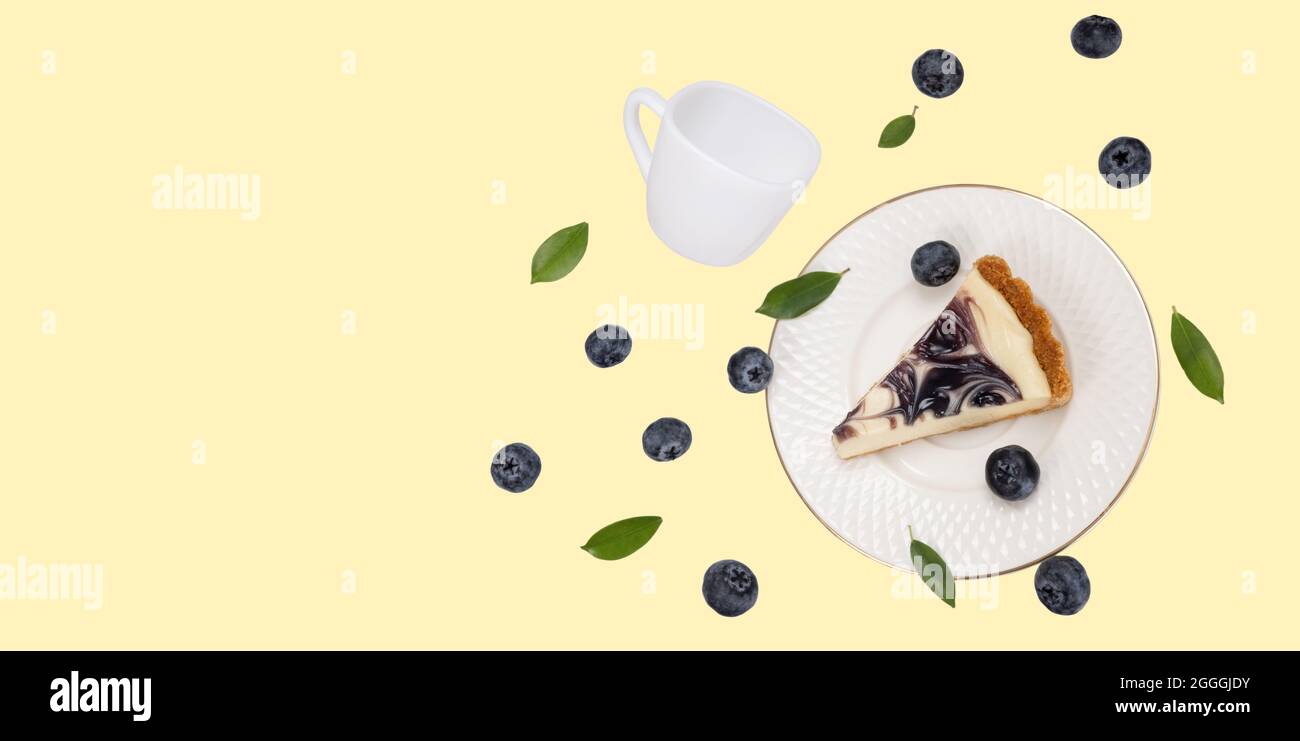 Ein Stück Heidelbeeren Käsekuchen auf einer fallenden weißen Keramikplatte mit fallenden weißen Keramikbecher und etwas Fruchtfleisch Blaubeeren und grünen Blättern auf weichen Stockfoto