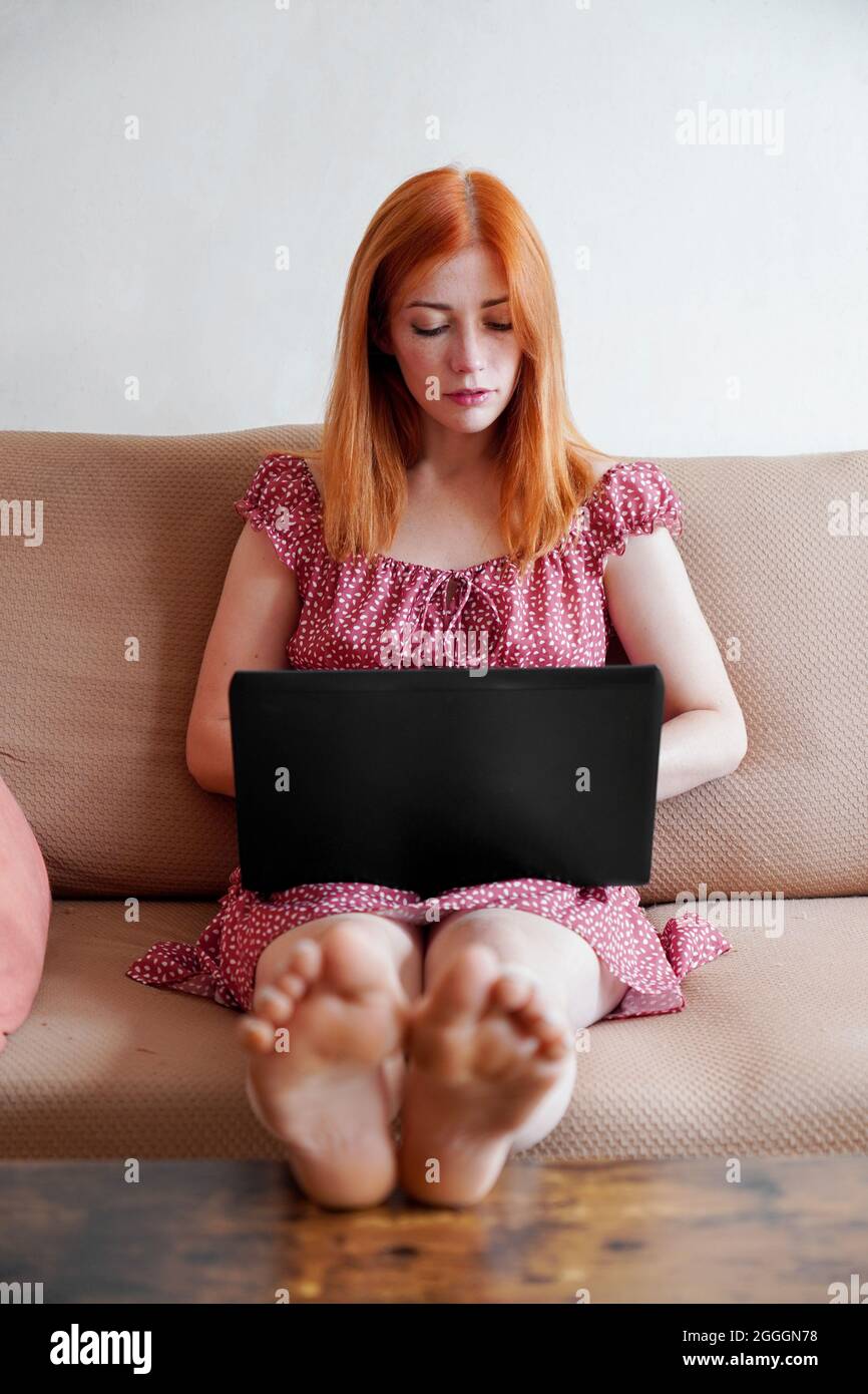 Junge Frau mit Laptop von zu Hause aus sitzend auf der Couch mit Füßen auf dem Tisch arbeiten Stockfoto
