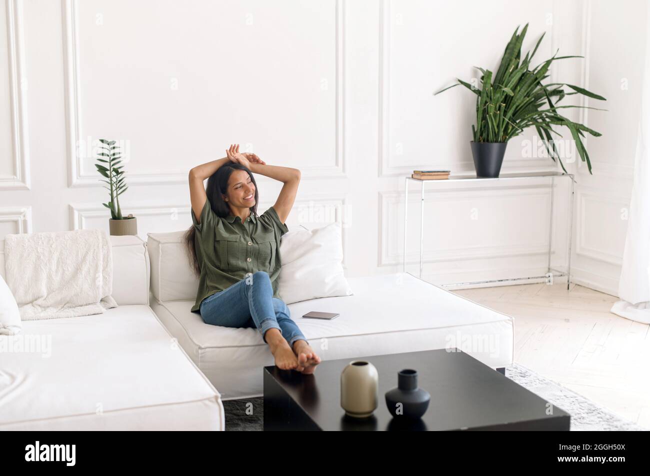 Harmonie und Balance Konzept. Unbeschwerte und gelassene indische Frau, die sich zu Hause auf der bequemen Couch ausruht, fühlt sich glücklich und heiter. Die charmante Dame aus dem Osten schaut weg und lächelt entspannt zu Hause Stockfoto