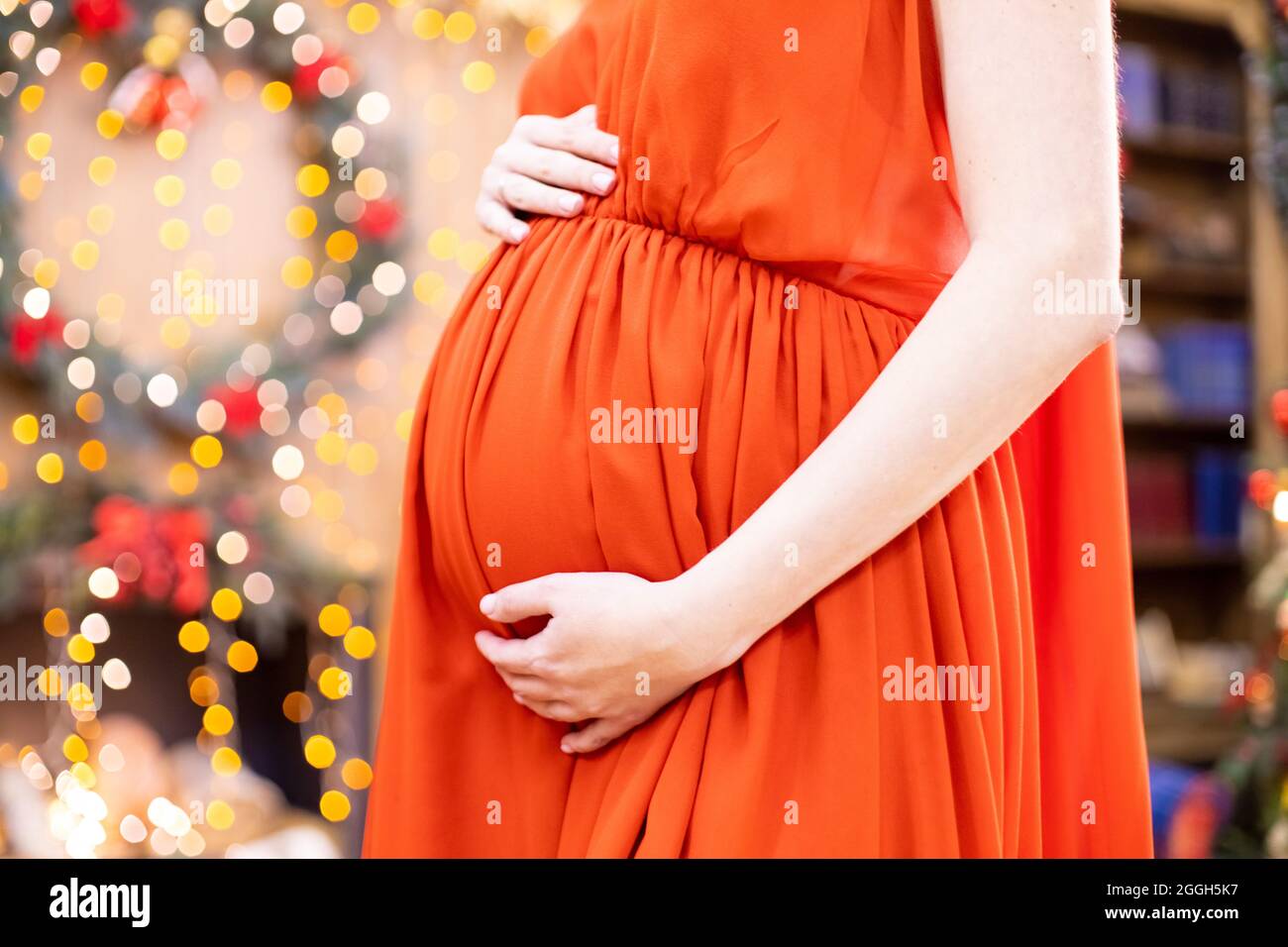 Bestes weihnachtsgeschenk. Gesichtslose Schwangere in einem roten Kleid umarmt ihren Bauch vor dem Hintergrund des Weihnachtswohnzimmers in Unschärfe. Nahaufnahme kein fa Stockfoto