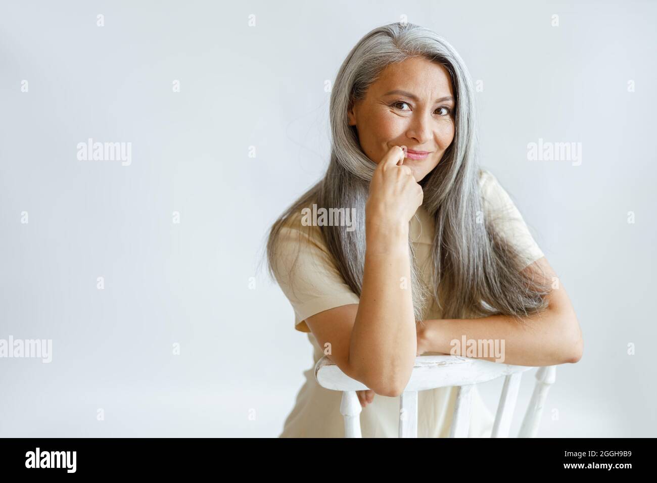 Flirten mittleren Alters asiatische Dame mit schönen grauen Haaren sitzt auf Stuhl auf hellem Hintergrund Stockfoto