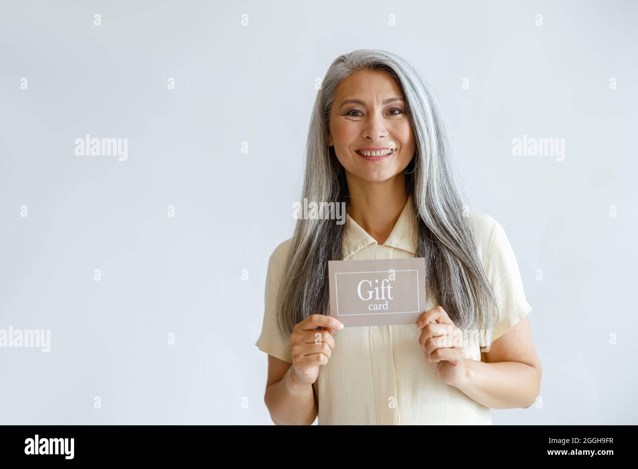 Lächelnde grauhaarige Asiatin hält den Geschenkgutschein auf hellem Hintergrund Stockfoto