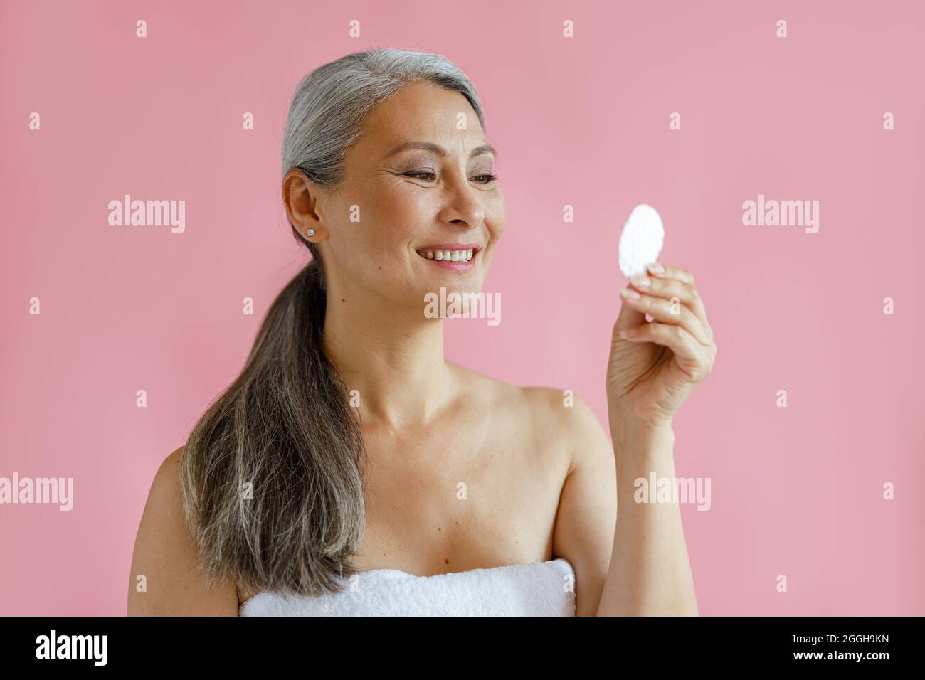 Schöne silberhaarige asiatische Frau sieht auf Baumwolle Pad stehen auf rosa Hintergrund Stockfoto