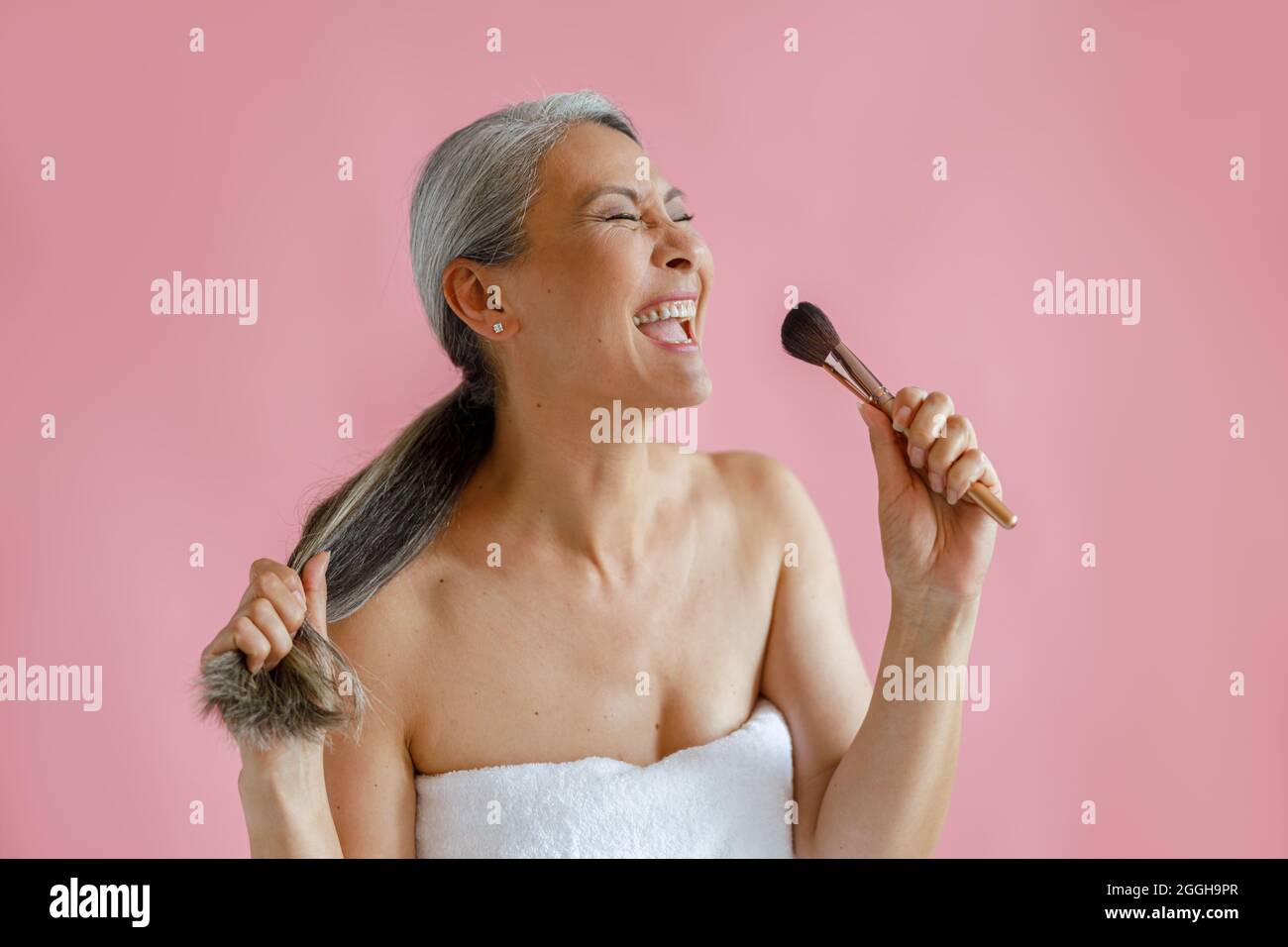 Jouful mittleren Alters asiatische Frau singt mit kosmetischen Pinsel als Mikrofon auf rosa Hintergrund Stockfoto