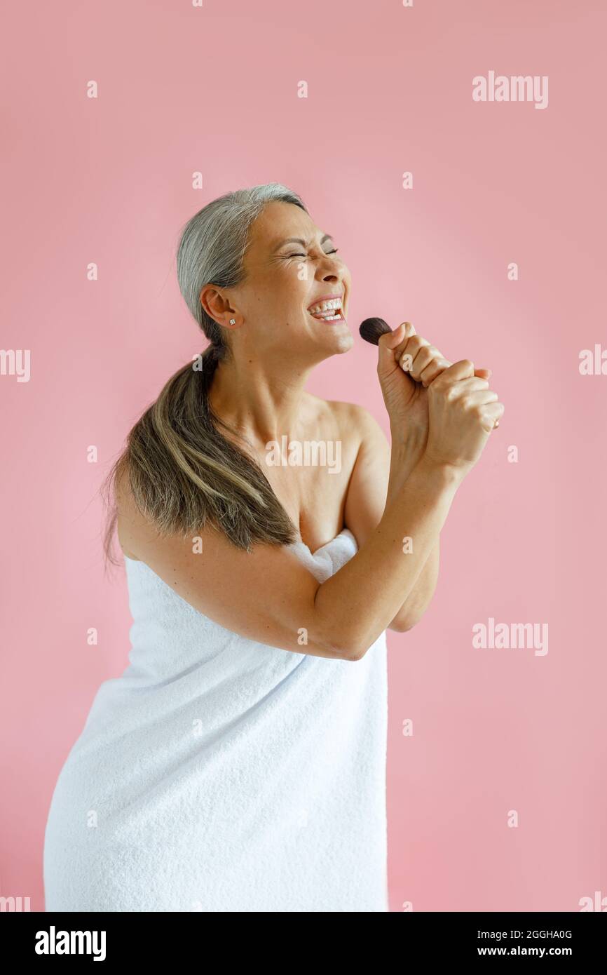 Glückliche silberhaarige asiatische Dame singt mit Pinsel stehen auf rosa Hintergrund Stockfoto
