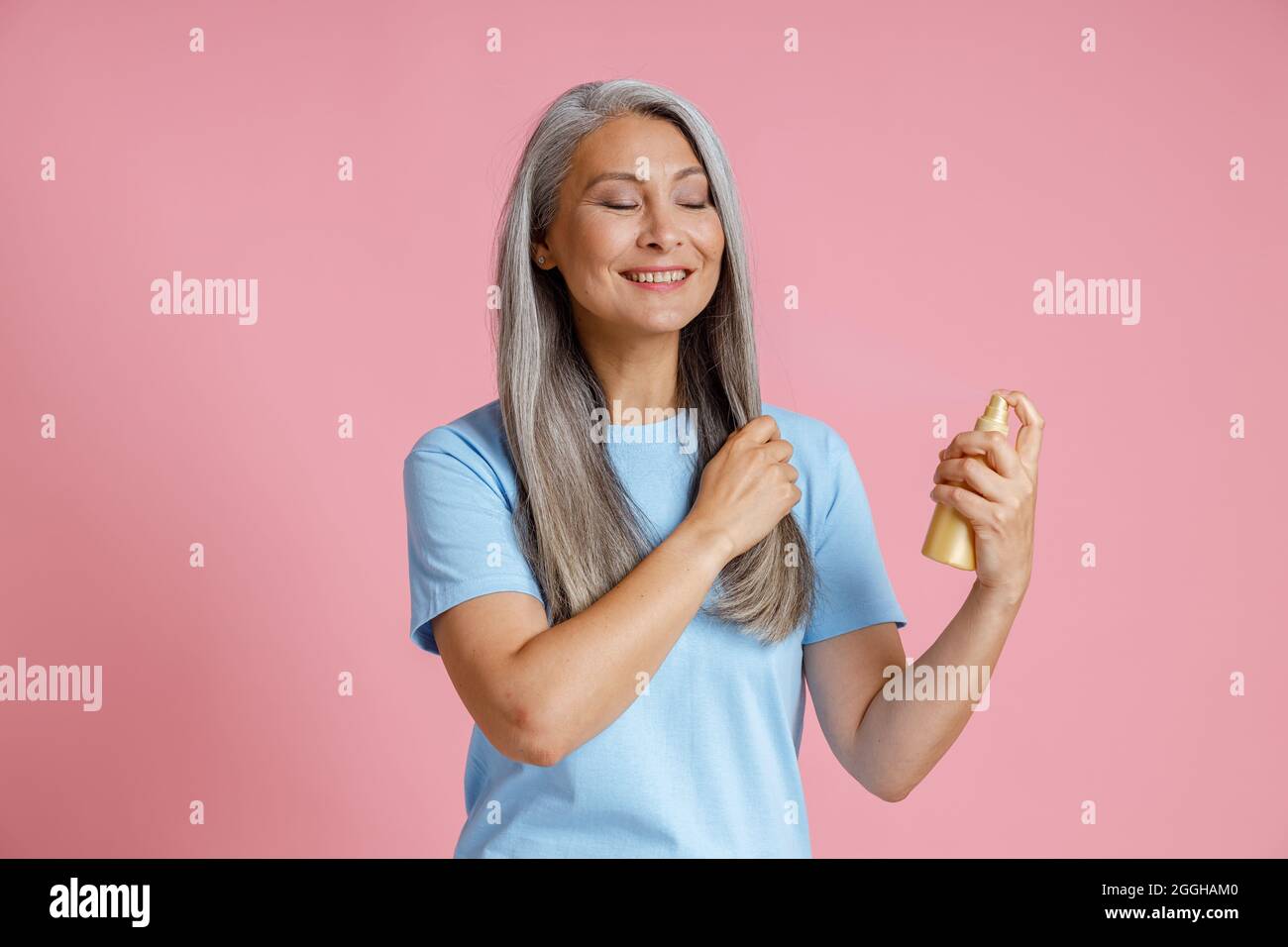 Positive reife asiatische Dame trägt Spray-Produkt auf lose graue Haare auf rosa Hintergrund Stockfoto