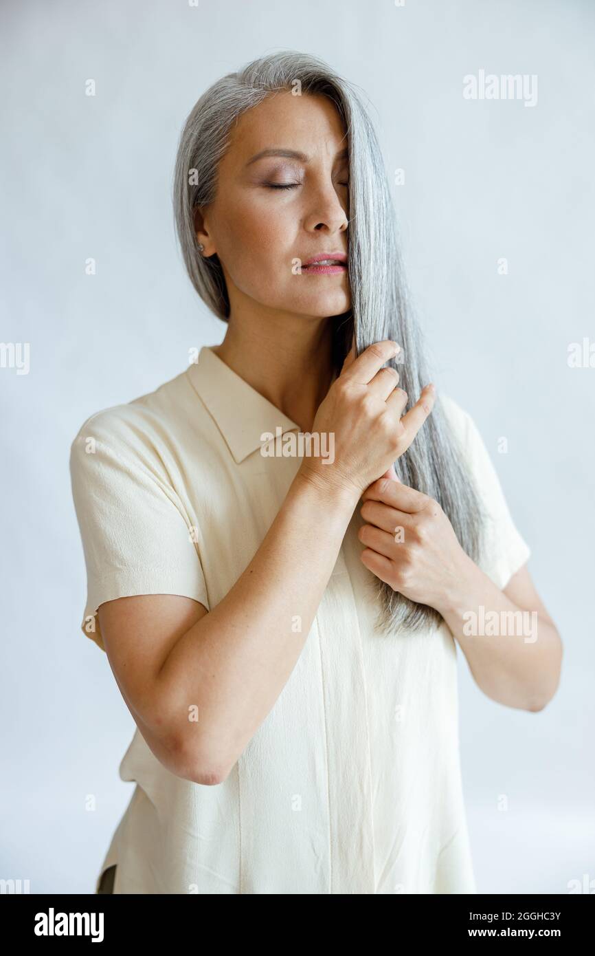 Languid mittleren Alters asiatische Frau streichelt Sperre der silbernen Haare auf hellgrauem Hintergrund Stockfoto