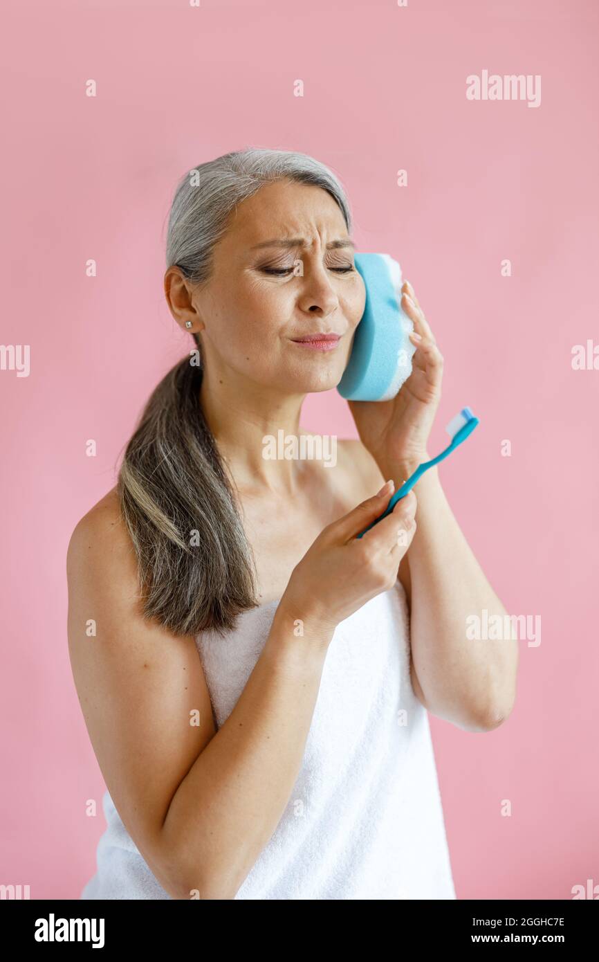 Eine ekelhafte Asiatin mittleren Alters sieht sich im Studio eine Zahnbürste an, die einen Badeschwamm im Ohr hält Stockfoto
