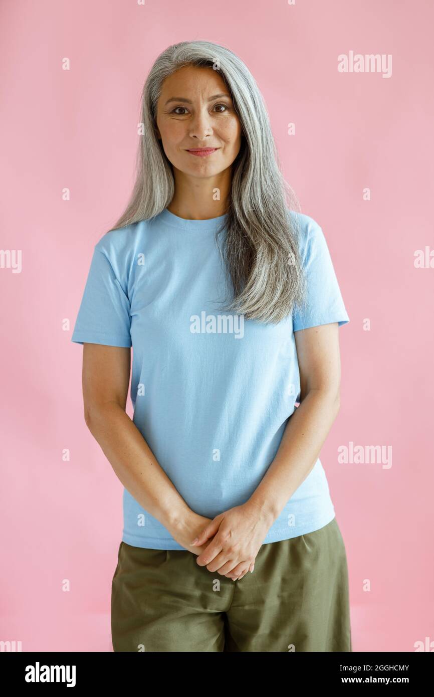 Ziemlich langhaarige asiatische Dame mittleren Alters in blauem T-Shirt steht auf rosa Hintergrund Stockfoto