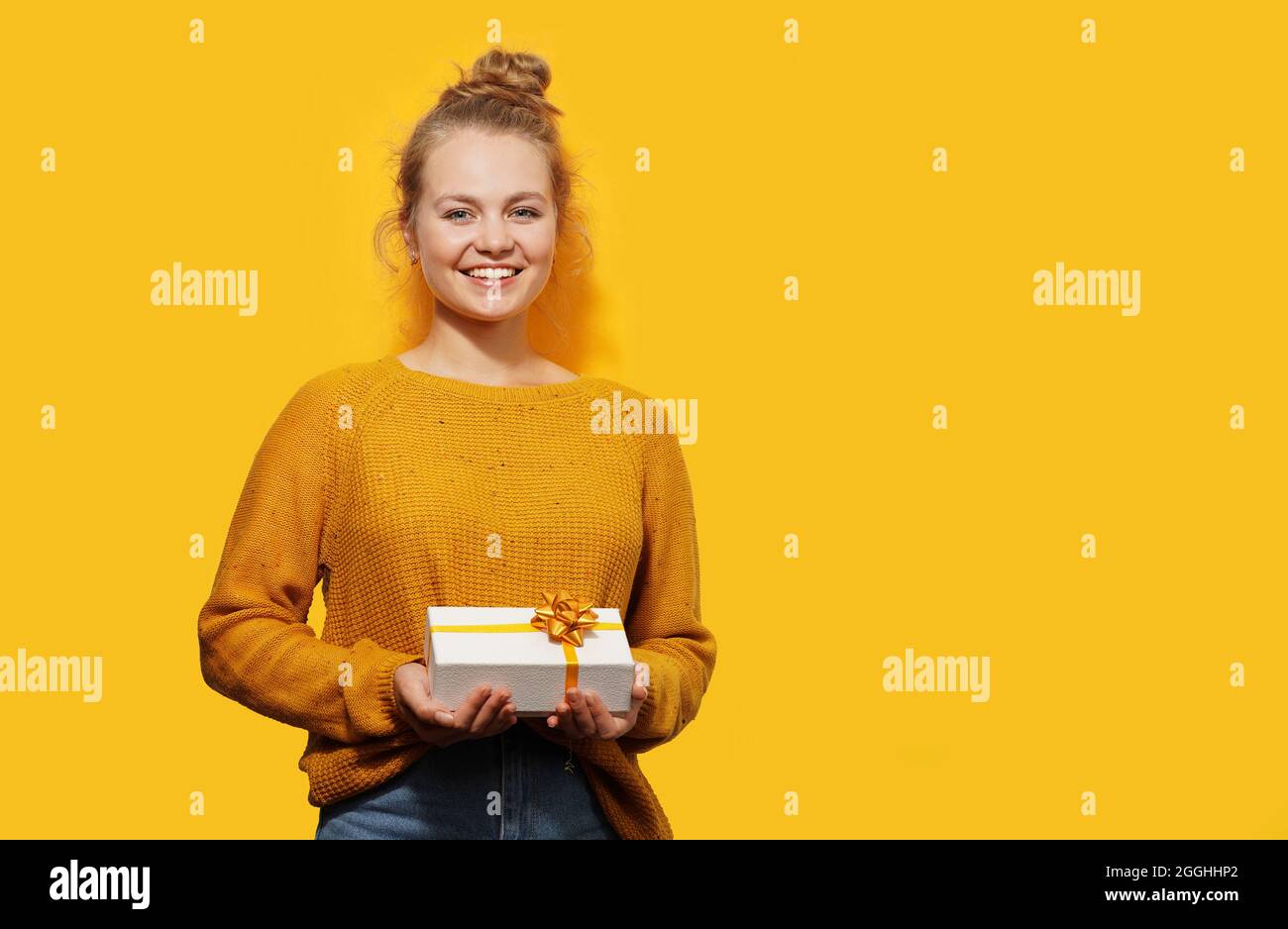 Lächelnd schöne blonde junge Frau hält eine Geschenkbox isoliert über gelbem Hintergrund. Alles gute zum Geburtstag, Weihnachten, Valentinstag-Konzept. Platz für Stockfoto