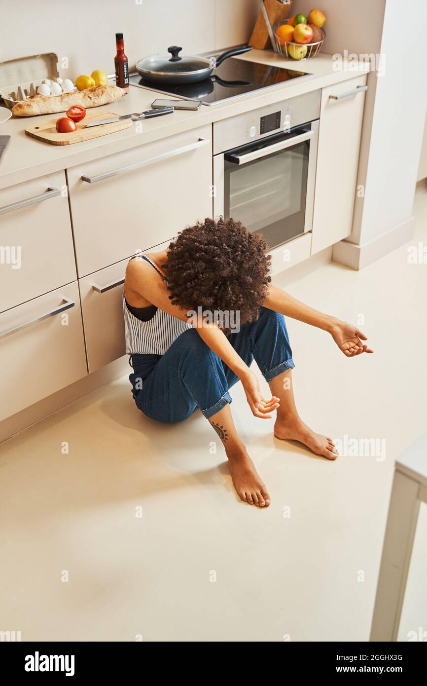Krestgefallene einsame, lockige junge afroamerikanische Frau, die barfuß auf dem Boden in der Küche sitzt Stockfoto
