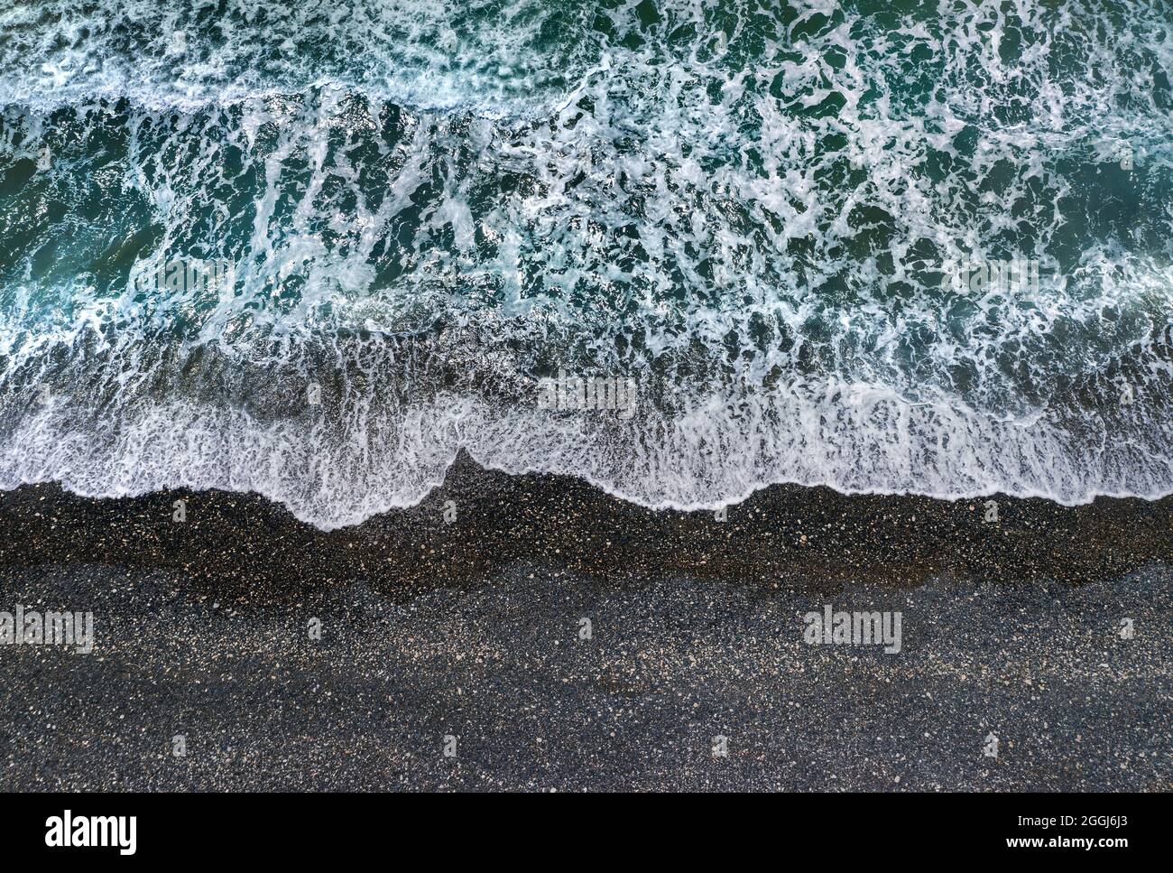 Stürmische grüne Wellen mit weißem Meeresschaum, der am dunklen Sandstrand bricht, Blick von direkt oben Stockfoto