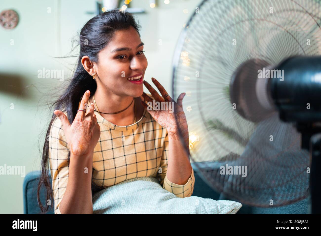 Glücklich lächelnde indische Frau, die während des Hitzeschlages auf der Couch sitzt, die Ventilatorluft genießt - Konzept der Hitzewelle oder das Gefühl der kühlen Luft aus dem Ventilator zu Hause. Stockfoto