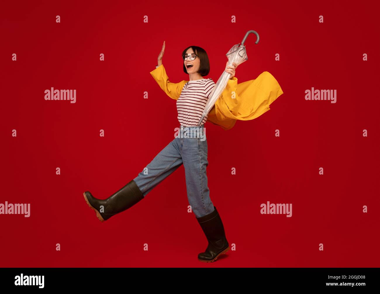 Herbstwetter. Junge Frau in Regenmantel und Gummistiefel kämpfen Wind und gehen über roten Hintergrund Stockfoto