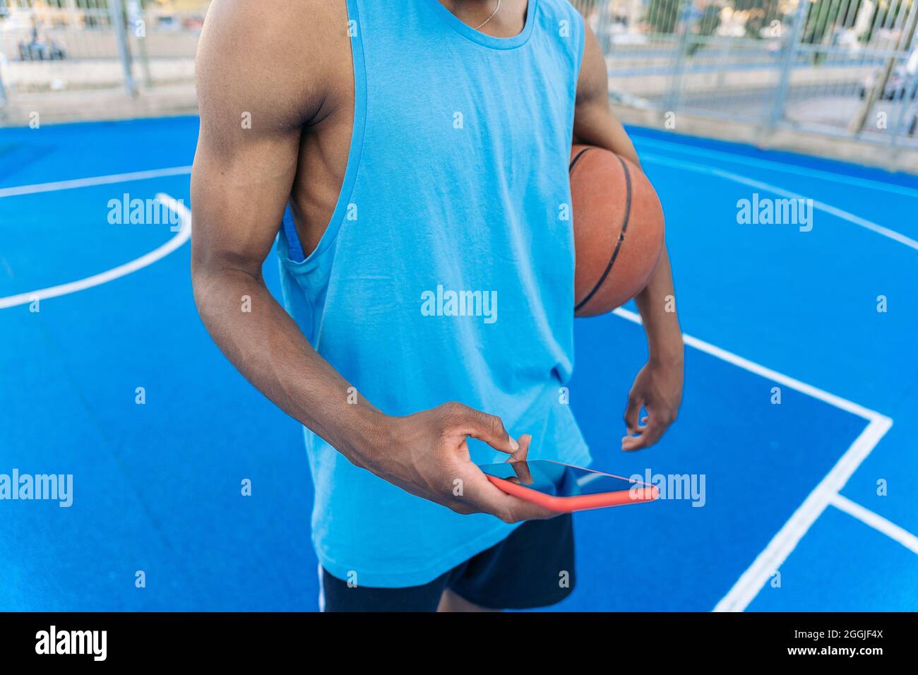 Junger afroamerikanischer Basketballspieler, der den Ball auf dem Basketballplatz der Straße unter seinen Arm hält, telefoniert. Stockfoto