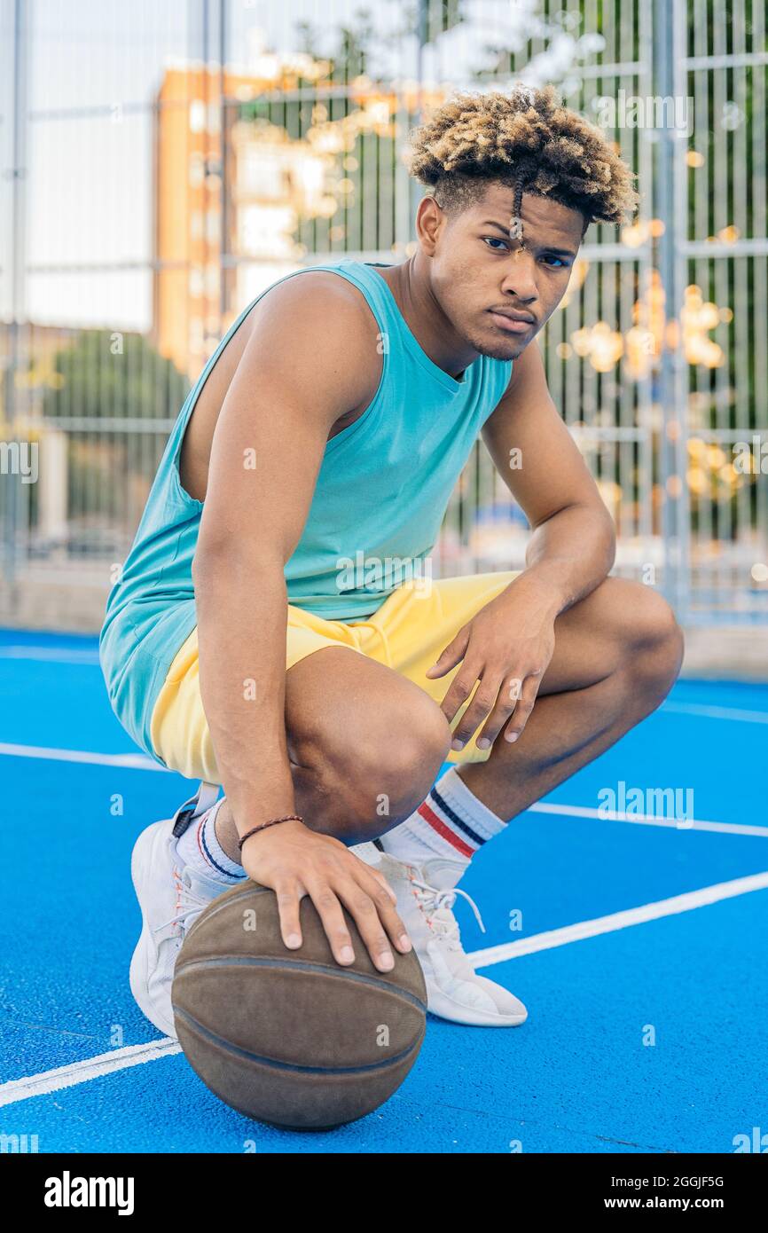 Porträt eines afro-amerikanischen Basketballspielers, der hockend den Ball auf dem Boden hält und ernsthaft die Kamera auf dem Platz anschaut. Stockfoto