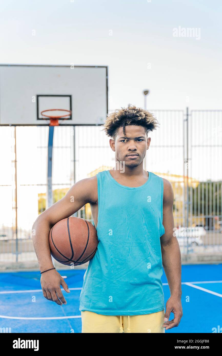 Ernsthafter afro-amerikanischer Basketballspieler, der die Kamera anschaut, die den Ball in einem Straßenbasketballfeld unter seinem Arm hält. Stockfoto
