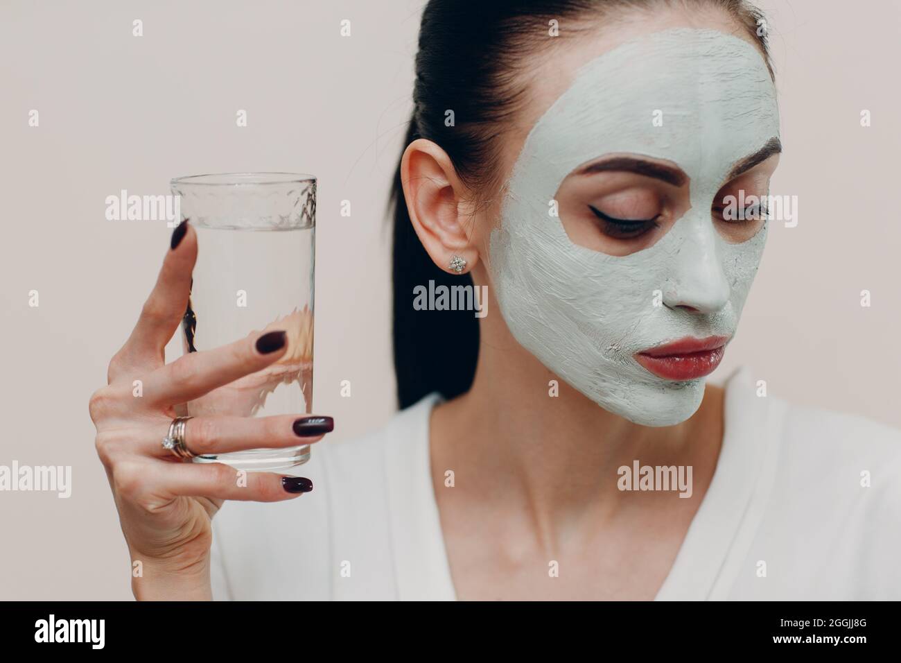 Junge schöne Frau erhält Gesichtsmaske aus Ton mit einem Glas Wasser. Stockfoto
