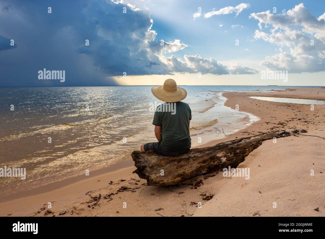 Mann im Hut sitzt auf Baumstamm und schaut auf Sturm und Sonnenuntergang am Tapajos-Flussstrand im Amazonas-Regenwald. Konzept von Natur, Naturschutz, Umwelt. Stockfoto