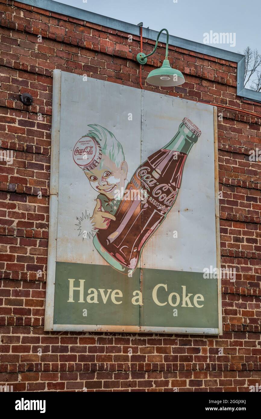 Ein altes, altes Coca-Cola-Werbeschild, das an einer Außenwand unter einem Licht auf einem Backsteingebäude angebracht war, das von einem jungen Holdi getragen und verblasst war Stockfoto