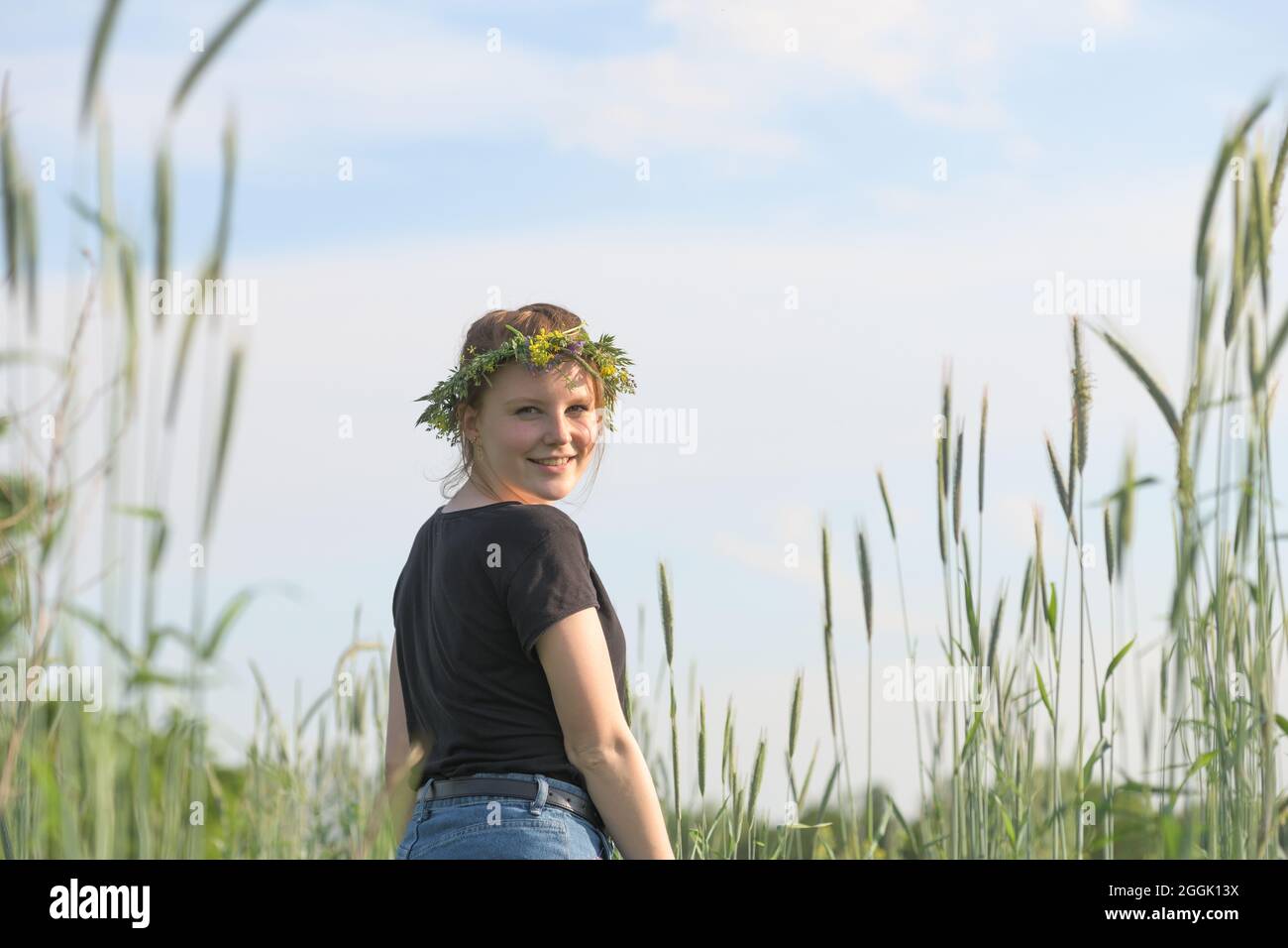 Die junge Frau mit einem Kranz aus natürlichen Blumen auf ihrem Kopf läuft lebhaft zwischen Getreidepflanzen Stockfoto