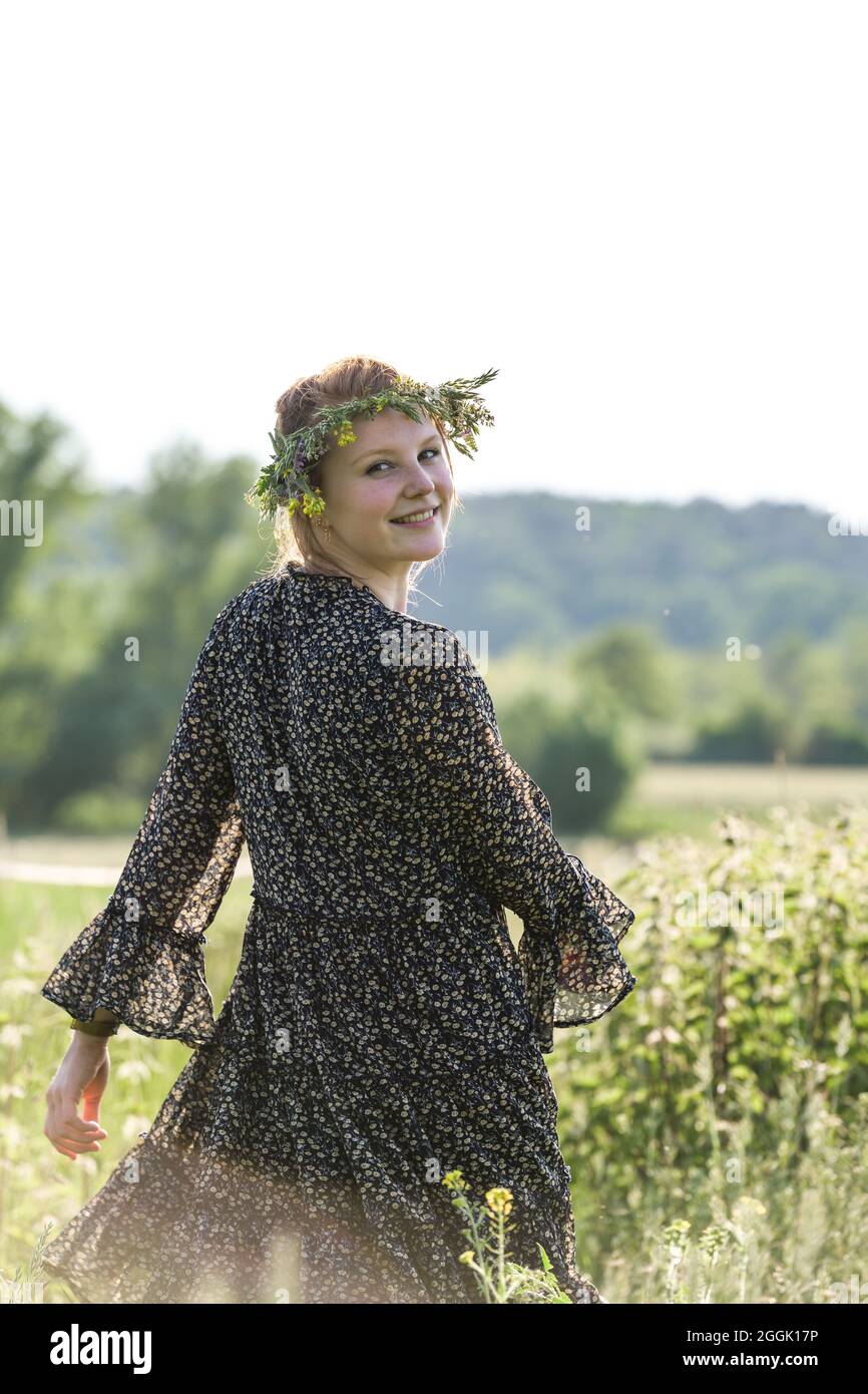 Die junge Frau mit einem Kranz natürlicher Blüten auf dem Kopf und Blumen in der Hand läuft lebhaft zwischen Feldern Stockfoto