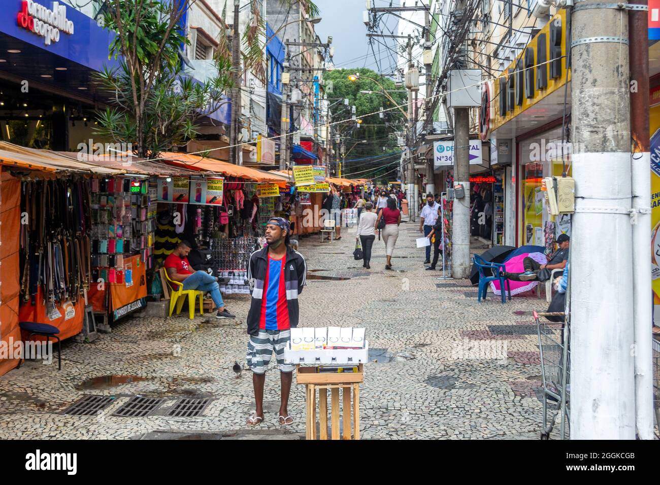 Ein brasilianischer Mann steht im Marktbezirk und versucht, Produkte mit einem kleinen tragbaren Tisch zu verkaufen. Andere Menschen werden im engen Bereich gesehen Stockfoto