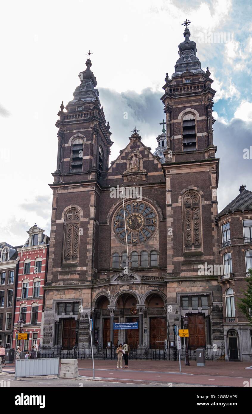 Amsterdam, Niederlande - 14. August 2021: Dunkelbraune monumentale Fassade mit Ornamenten, Statue und 2 Türmen der Sankt-Nikolaus-Basilika unter grauer Wolke Stockfoto