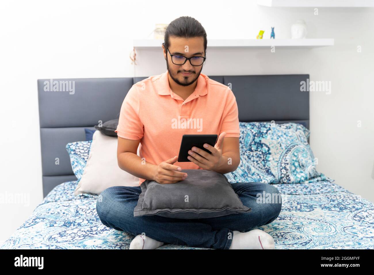 Junger Mann, der auf dem Bett sitzt und auf einem E-Reader liest Stockfoto