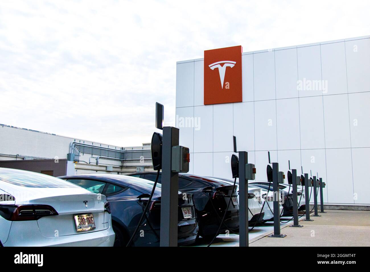 Tesla-Logo auf der Seite eines Gebäudes, hinter einer Reihe von charing Tesla Model 3 und Y, bereit, vom Schaufenster aus getestet zu werden. Stockfoto