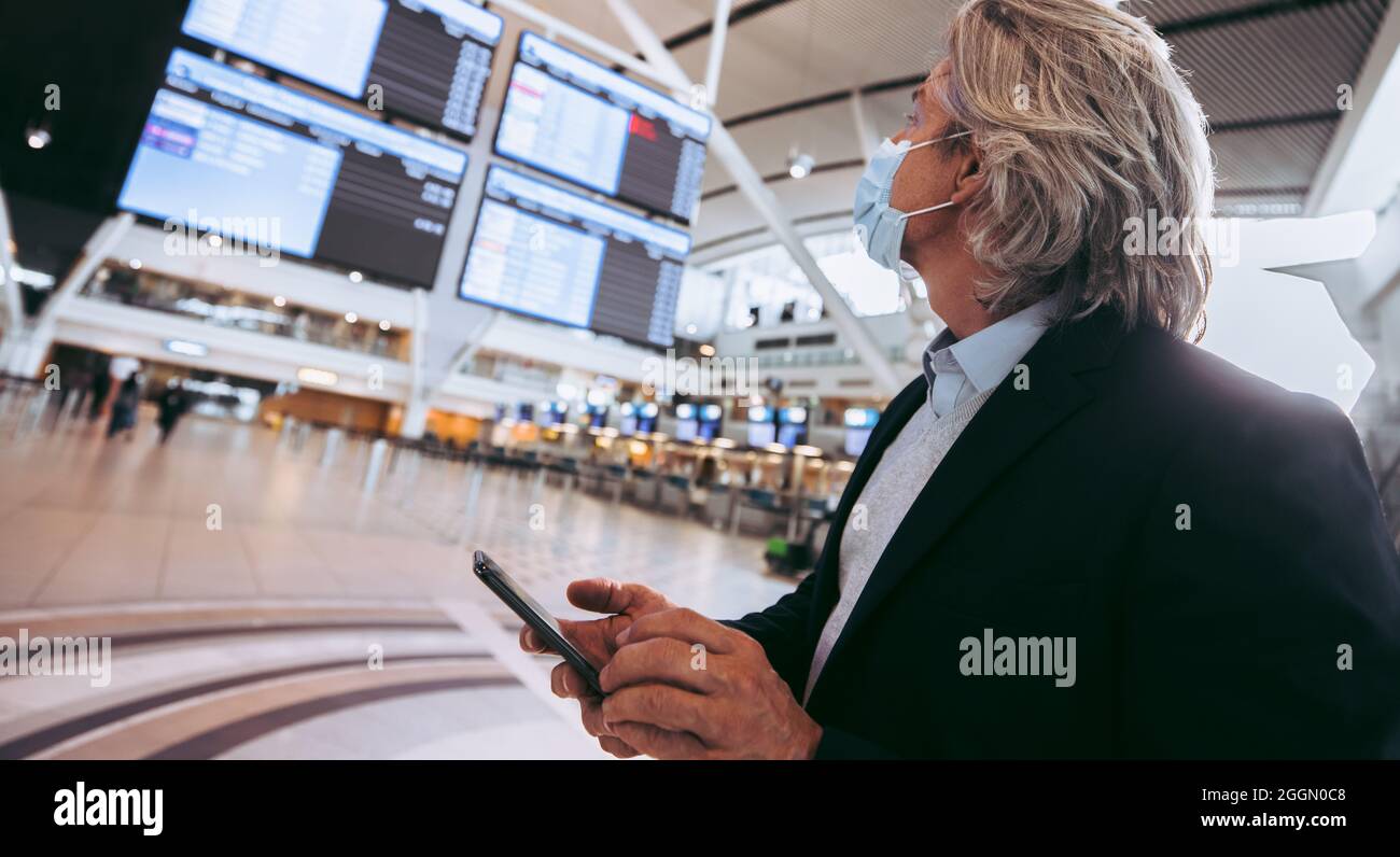Geschäftsmann mit Gesichtsmaske und Blick auf das Abflugbrett im Flughafenterminal. Männlicher Reisender, der auf dem Bildschirm „Fluginformationen“ im Flughafen zusieht Stockfoto