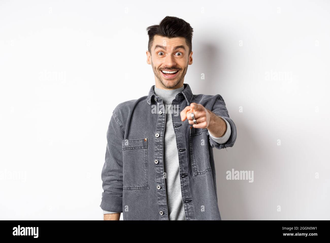 Du bist es. Aufgeregter lächelnder Mann, der mit fröhlichem Gesicht mit den Fingern auf die Kamera zeigt, erkennt die Person, die in einer Jeansjacke auf weißem Hintergrund steht. Stockfoto
