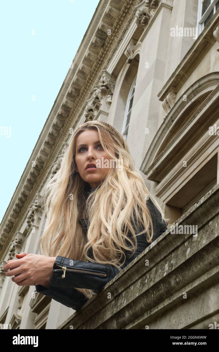 Blick auf eine blonde Frau, die sich über den Balkon eines alten, kunstvoll verzierten Gebäudes lehnt. Stockfoto