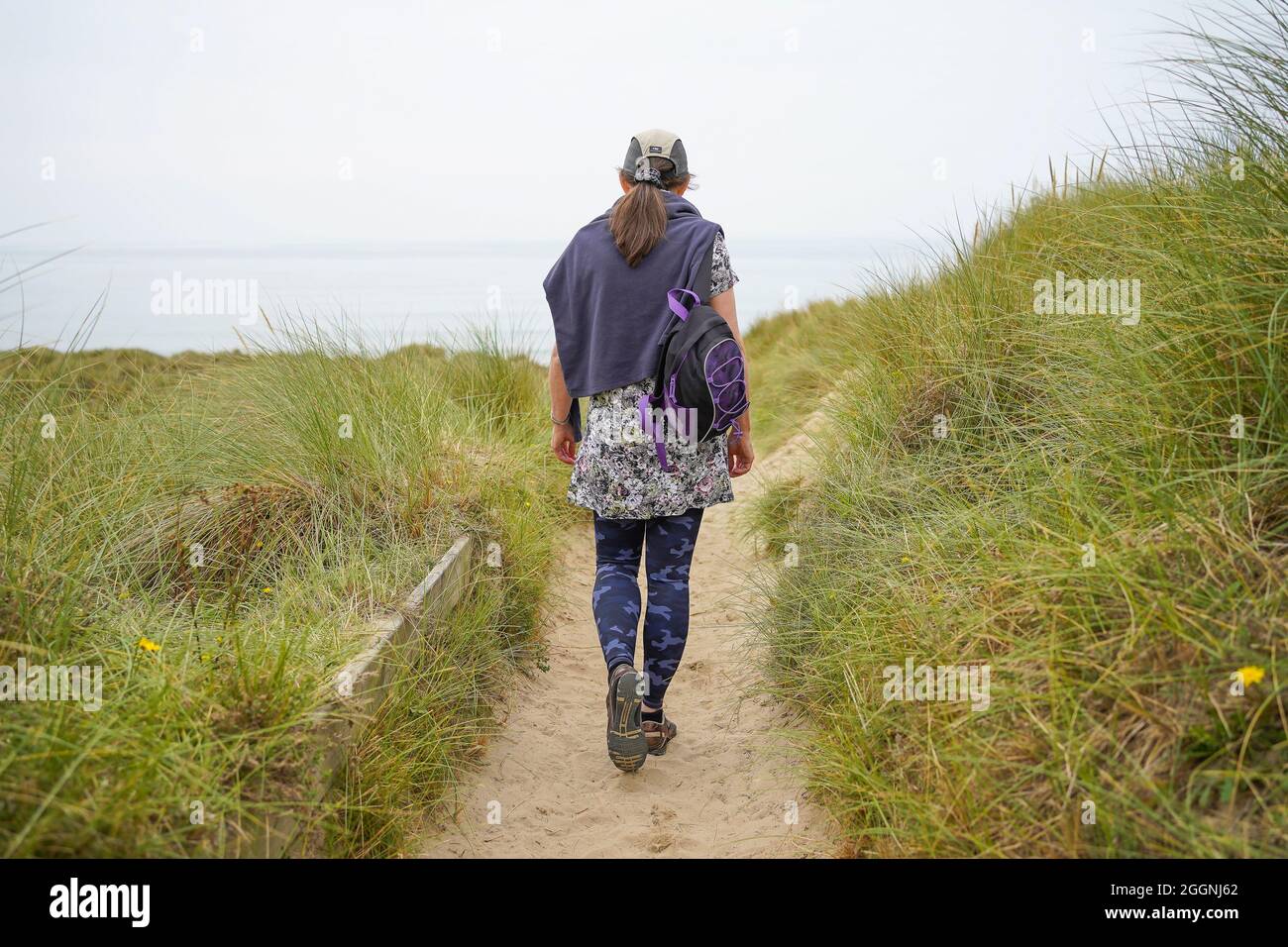 Rückansicht einer jungen Frau, die an einem bewölkten Tag in Wales in Richtung Strand und Meer vor ihr geht Stockfoto