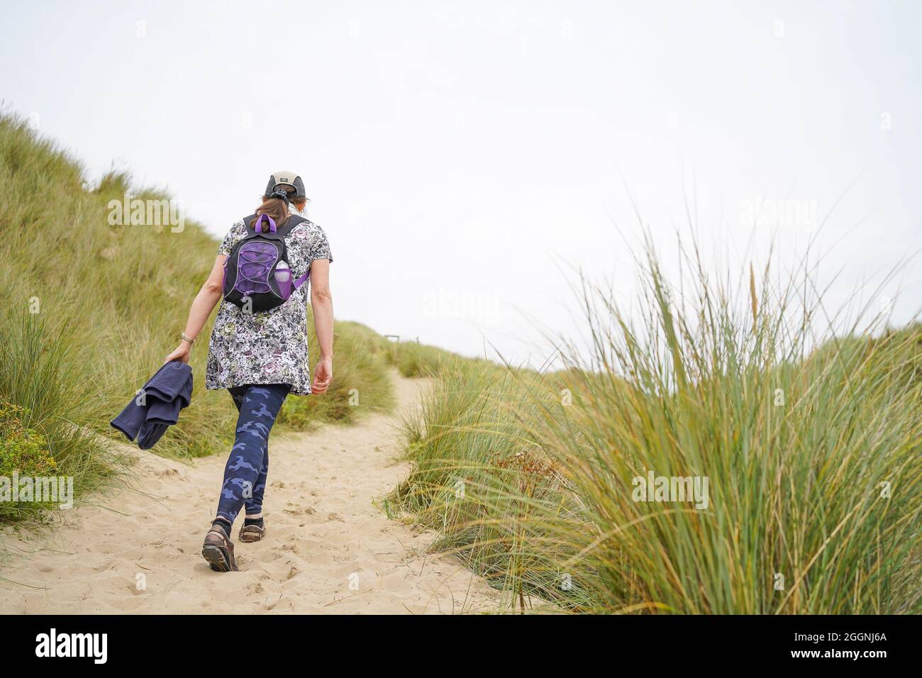 Rückansicht einer isolierten jungen Frau, die an einem bewölkten Sommertag in Wales, Großbritannien, in Küstensanddünen zur Strandpromenade läuft. Stockfoto