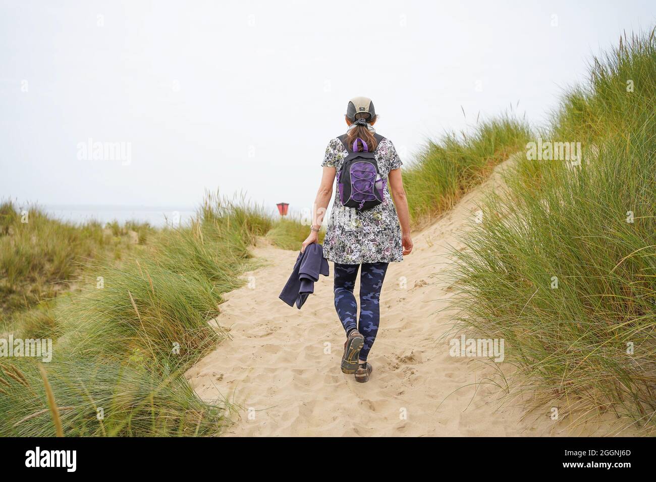 Rückansicht einer isolierten jungen Frau in Sportmütze, die an einem bewölkten Sommertag in Wales in Küstensanddünen in Richtung Strandpromenade geht. Stockfoto