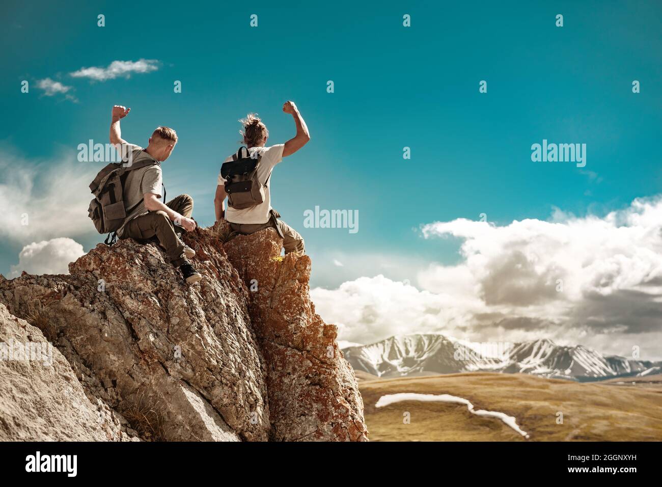Zwei starke Wanderer mit Rucksäcken sitzen auf einem großen Felsen in Siegerpose. Konzept für Bergurlaube Stockfoto