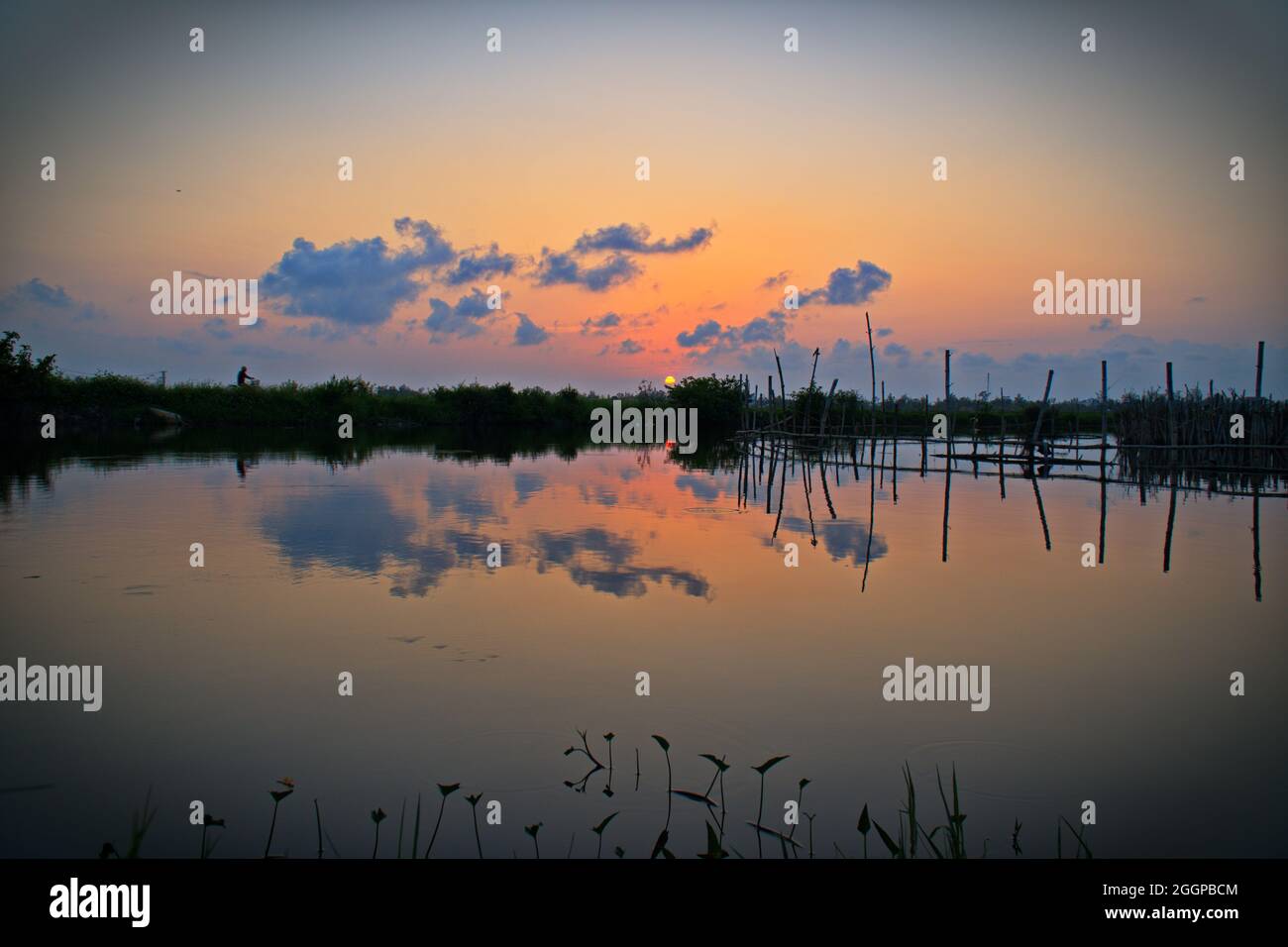 Friedlicher Sonnenuntergang über einem ruhigen Feuchtgebiet mit Fischernetzen und Silhouetten-Vegetation, die leuchtende Orange-, Rosa- und Blautöne im stillen Wasser reflektiert. Stockfoto