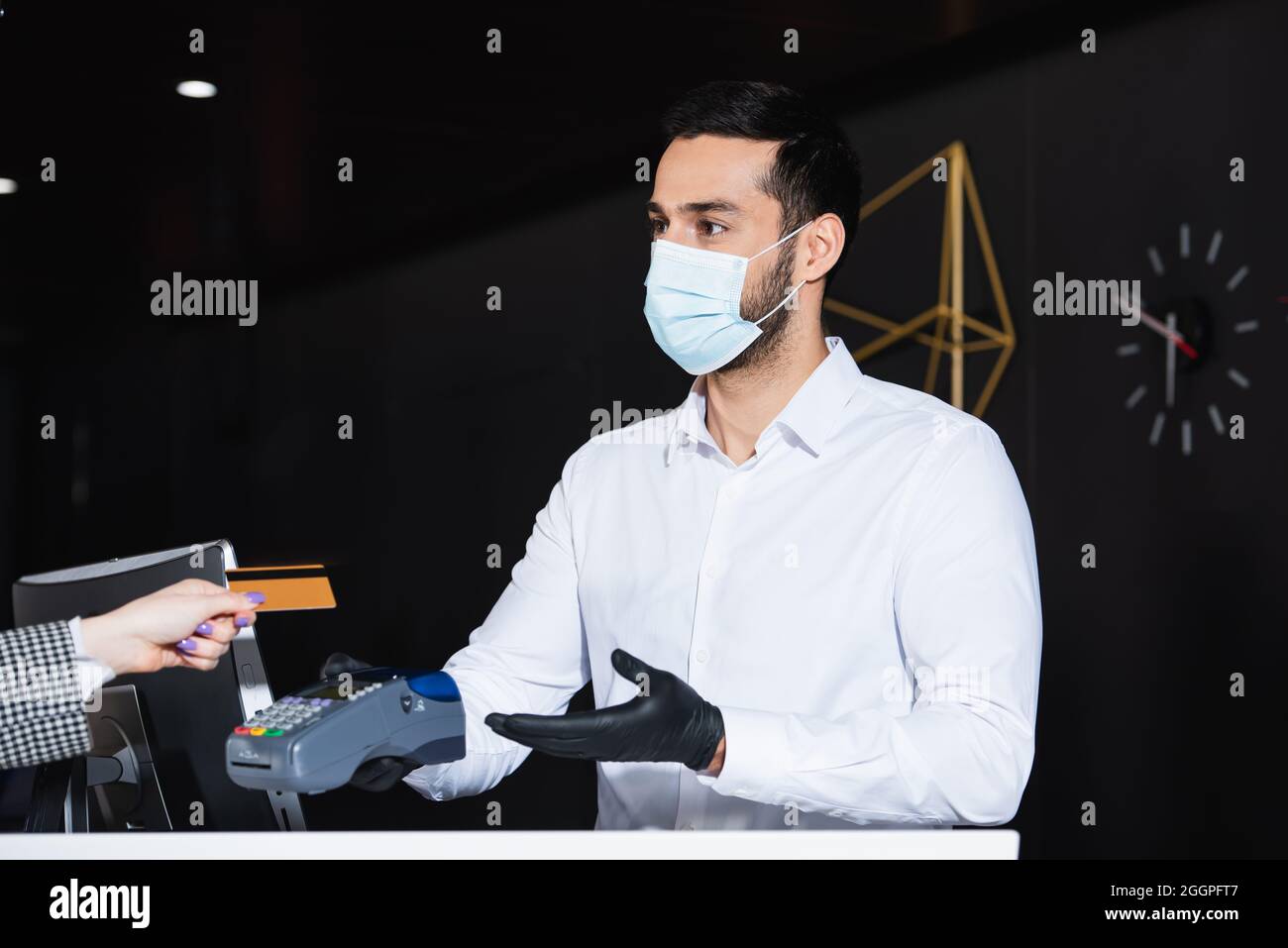 Rezeptionist mit medizinischer Maske und Latexhandschuhen, die das Zahlungsterminal mit Kreditkarte in der Nähe des Touristen halten Stockfoto