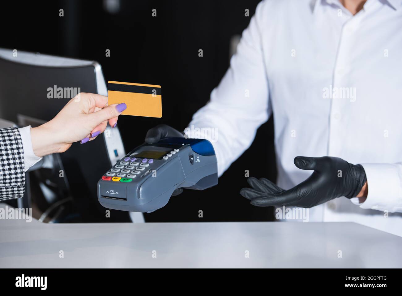 Abgeschnittene Ansicht der Empfangsdame in Latexhandschuhen, die das Zahlungsterminal mit Kreditkarte in der Nähe des Touristen halten Stockfoto