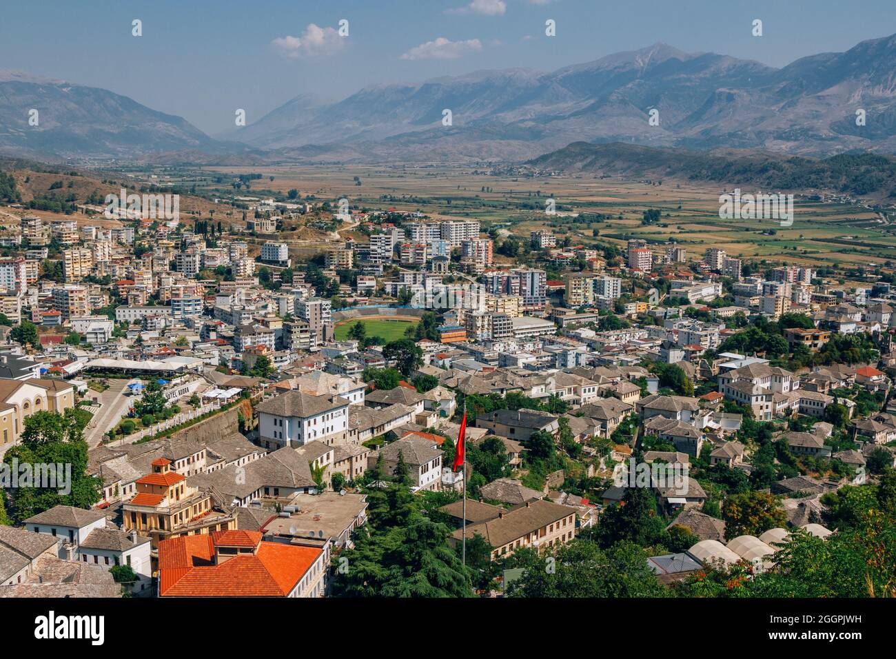 Stadtbild Gjirokastra – traditionelle albanische weiße Häuser mit steinernen grauen Dächern und modernen Gebäuden. Sommerlandschaft mit üppigem Laub, blauem Himmel und Stockfoto