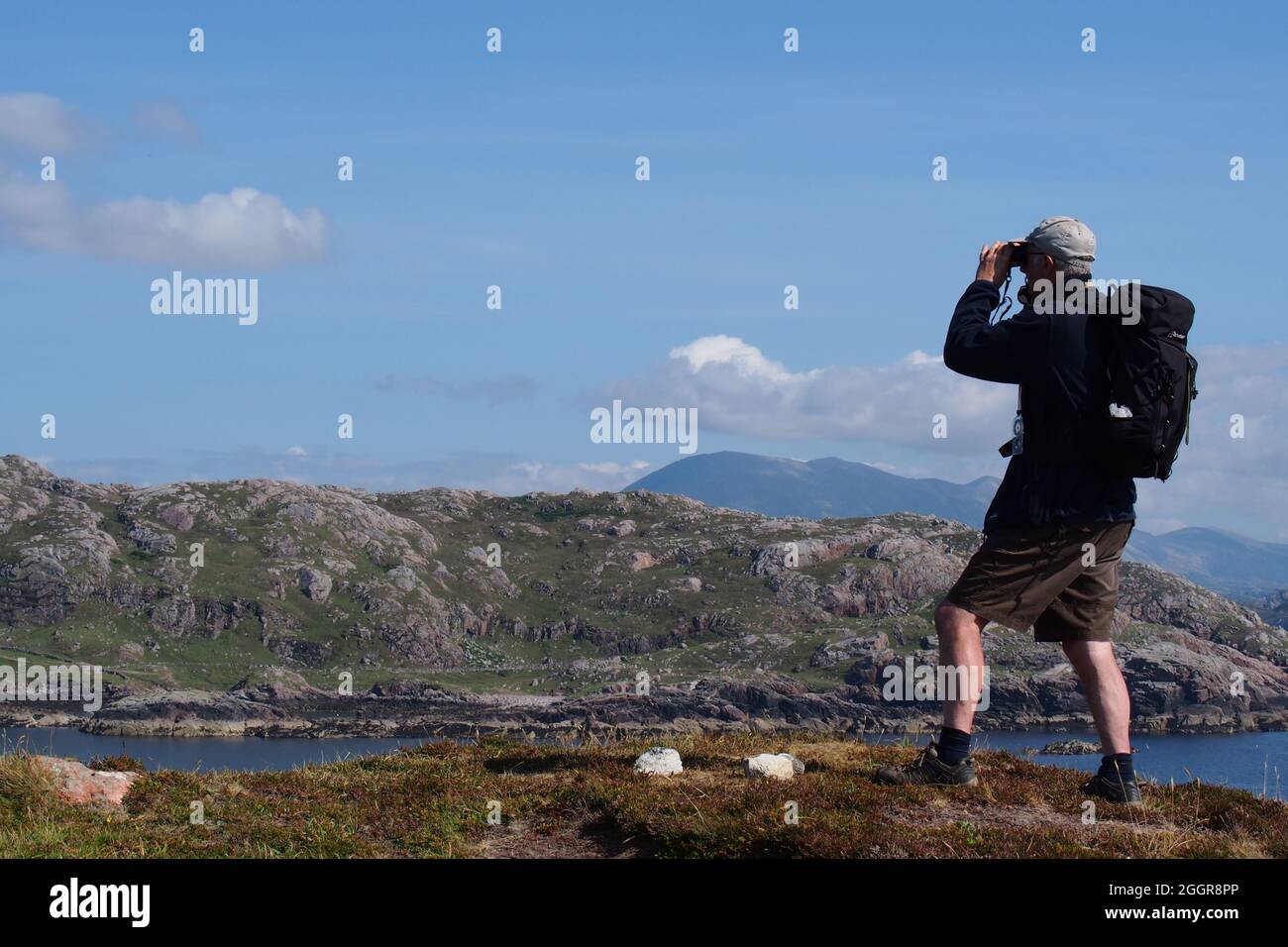 Ein über 60 Mann, der einen Rucksack mit Mütze und Shorts trägt und durch ein Fernglas über eine Meereslandschaft bis zu Land und Bergen in Schottland blickt, bleibt hier Stockfoto