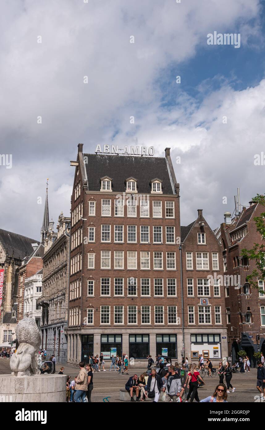 Amsterdam, Niederlande - 14. August 2021: Brauner Stein mit dunkler Dachecke Gebäude der ABN-AMRO Bank am Dam unter blauer Wolkenlandschaft. Viele Leute c Stockfoto